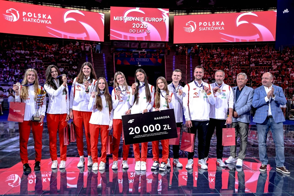 Polskie mistrzynie Europy U-16 uhonorowane w czasie finałów Ligi Narodów siatkarek Grupa siatkarek i trenerów pozuje na parkiecie hali sportowej z dużym symbolicznym czekiem na 20 000 zł, medalami oraz upominkami na tle pełnej trybuny kibiców i ekranów prezentujących napisy o mistrzostwie Europy do lat 17 w siatkówce.
