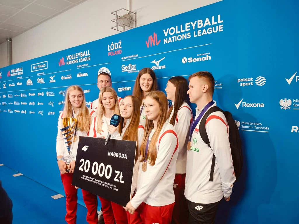 Polskie mistrzynie Europy siatkarek U-16 w czasie finałów Ligi Narodów Grupa młodych zawodniczek i zawodników w biało-czerwonych strojach sportowych pozuje do zdjęcia na tle ścianki promującej Volleyball Nations League, trzymając wielki symboliczny czek na 20 000 zł nagrody.