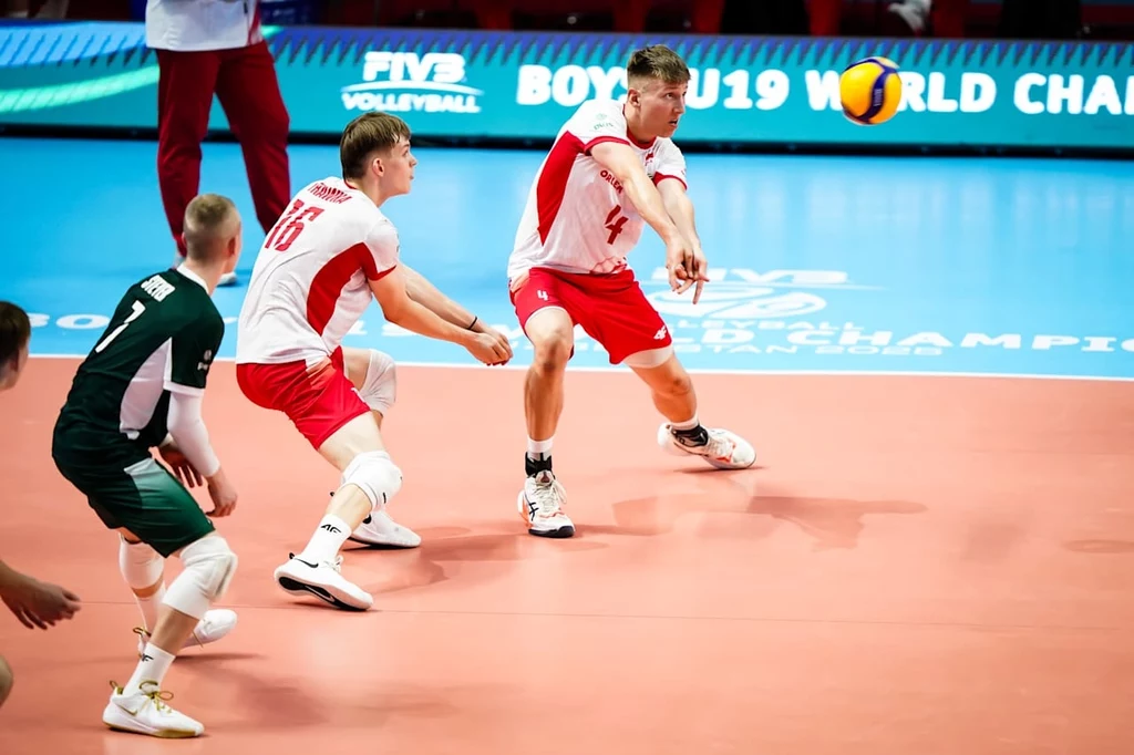 Siatkarze reprezentacji Polski podczas meczu z Hiszpanią na MŚ U19 Dwóch polskich siatkarzy w białych i czerwonych strojach wykonuje odbiór piłki podczas meczu, obok widać zawodnika przeciwnej drużyny w zielonym stroju, tło stanowi niebieskie boisko z napisem FIVB Boys' U19 World Championship.