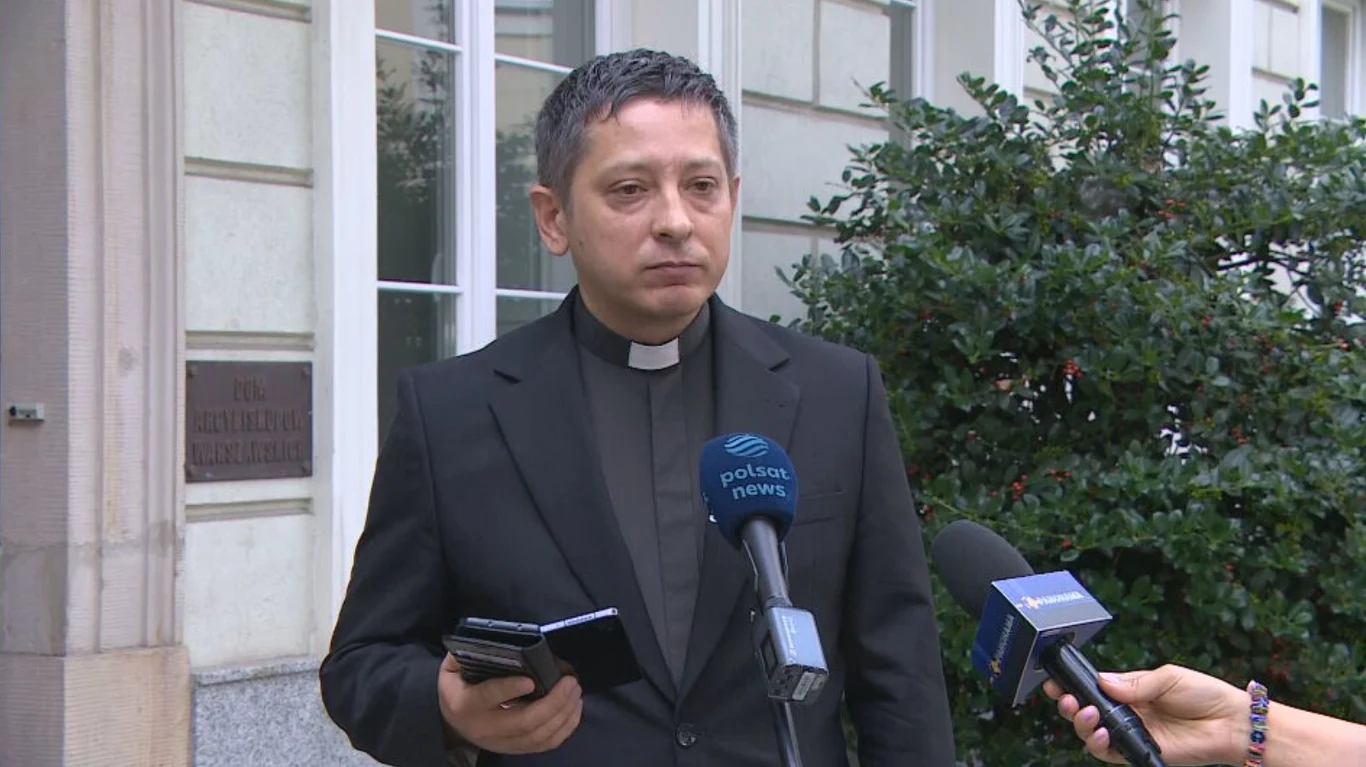 Kuria warszawska reaguje po zarzutach dla proboszcza Mężczyzna w koloratce trzymający telefon oraz notes, udzielający wywiadu przed budynkiem, otoczony przez mikrofony dziennikarzy.