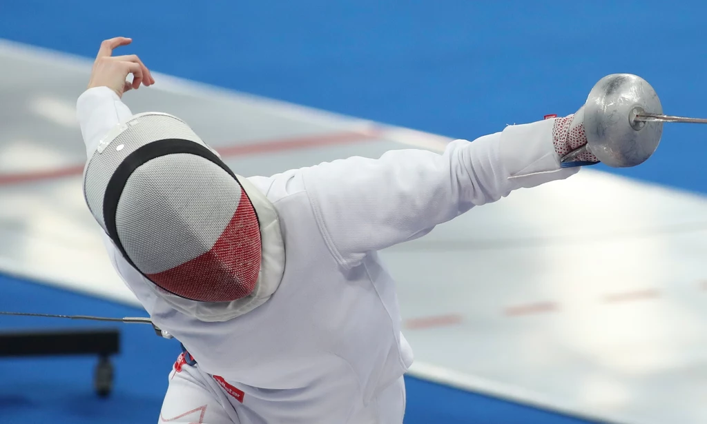 Alicja Klasik Szermierz w białym stroju i masce wykonuje dynamiczny atak szpadą na niebieskiej planszy sportowej.