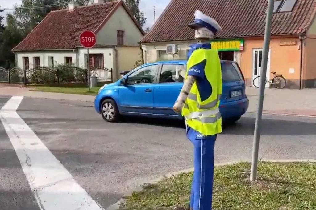 Manekin ubrany w strój policyjny stoi przy skrzyżowaniu, obok niego przejeżdża niebieski samochód, w tle widoczne są budynki z czerwoną dachówką oraz znak STOP.