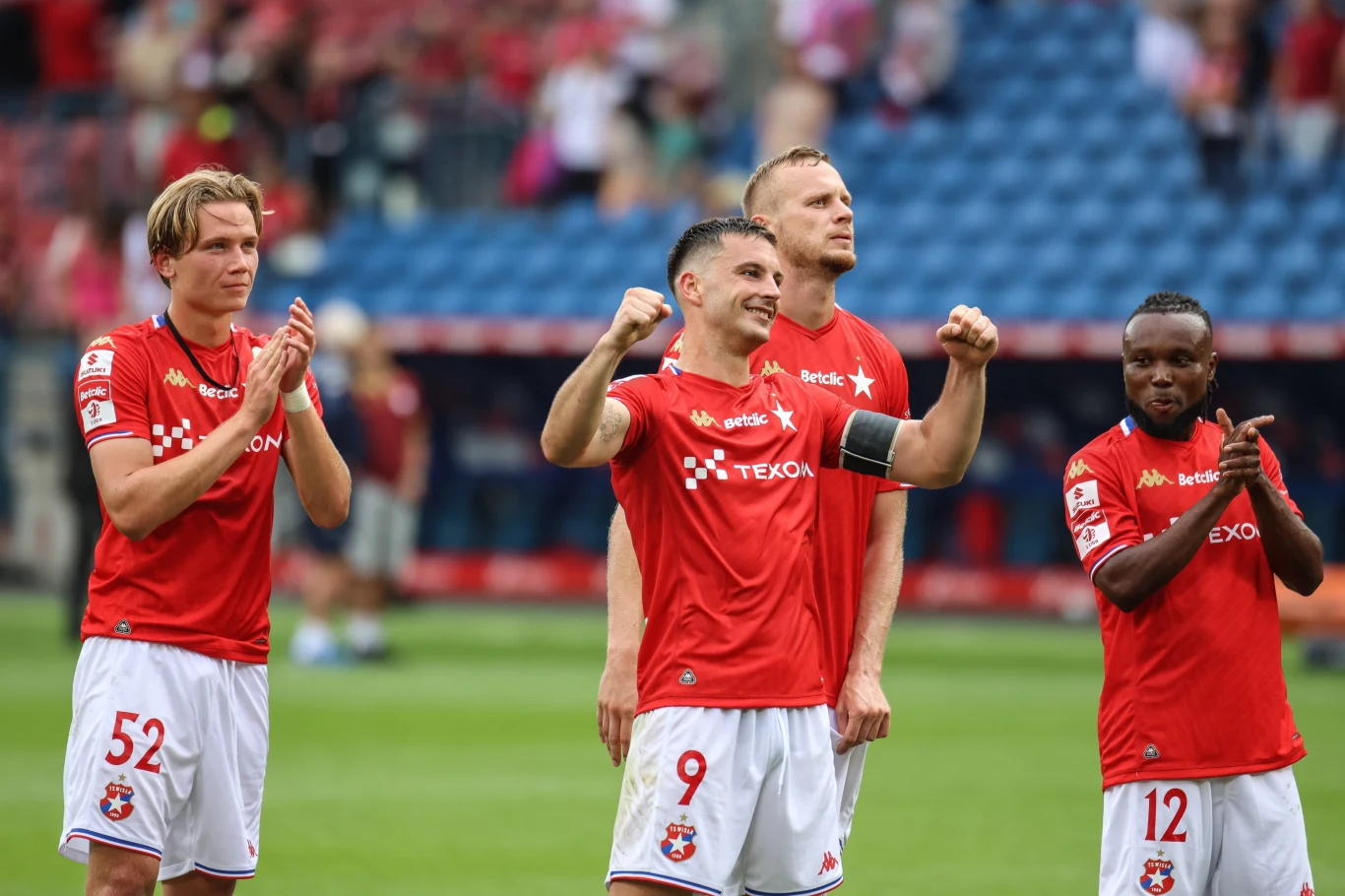 Wisła Kraków Czterech piłkarzy drużyny ubranych w czerwone koszulki stoi na boisku, trzech z nich bije brawo, a jeden zawodnik z opaską kapitana wznosi ręce w geście triumfu; w tle widzowie na trybunach.