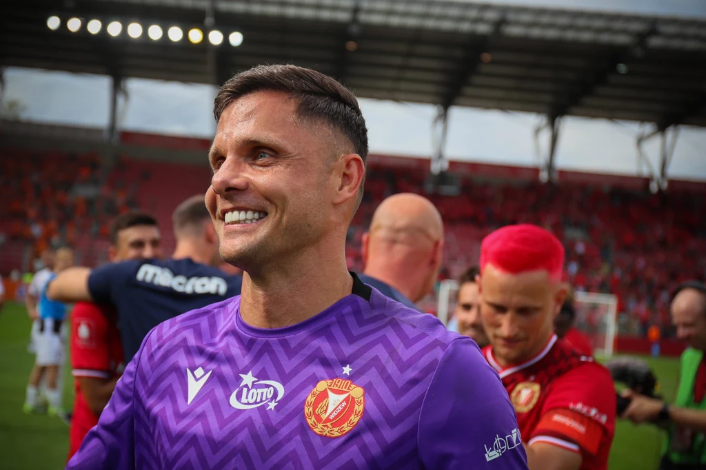 Piłkarz w fioletowej koszulce łódzkiego klubu z uśmiechem na twarzy na tle stadionu, kibice i członkowie drużyny w tle, w tym jedna osoba z intensywnie różowymi włosami, atmosfera celebracji po meczu.
