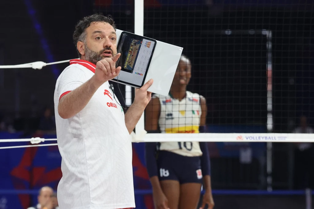 Stefano Lavarini w trakcie meczu Polska - Włochy w półfinale Ligi Narodów Trener w białej koszulce z tabletem w ręku wydaje instrukcje podczas meczu siatkówki; w tle siatkarka w stroju sportowym stoi przy siatce.