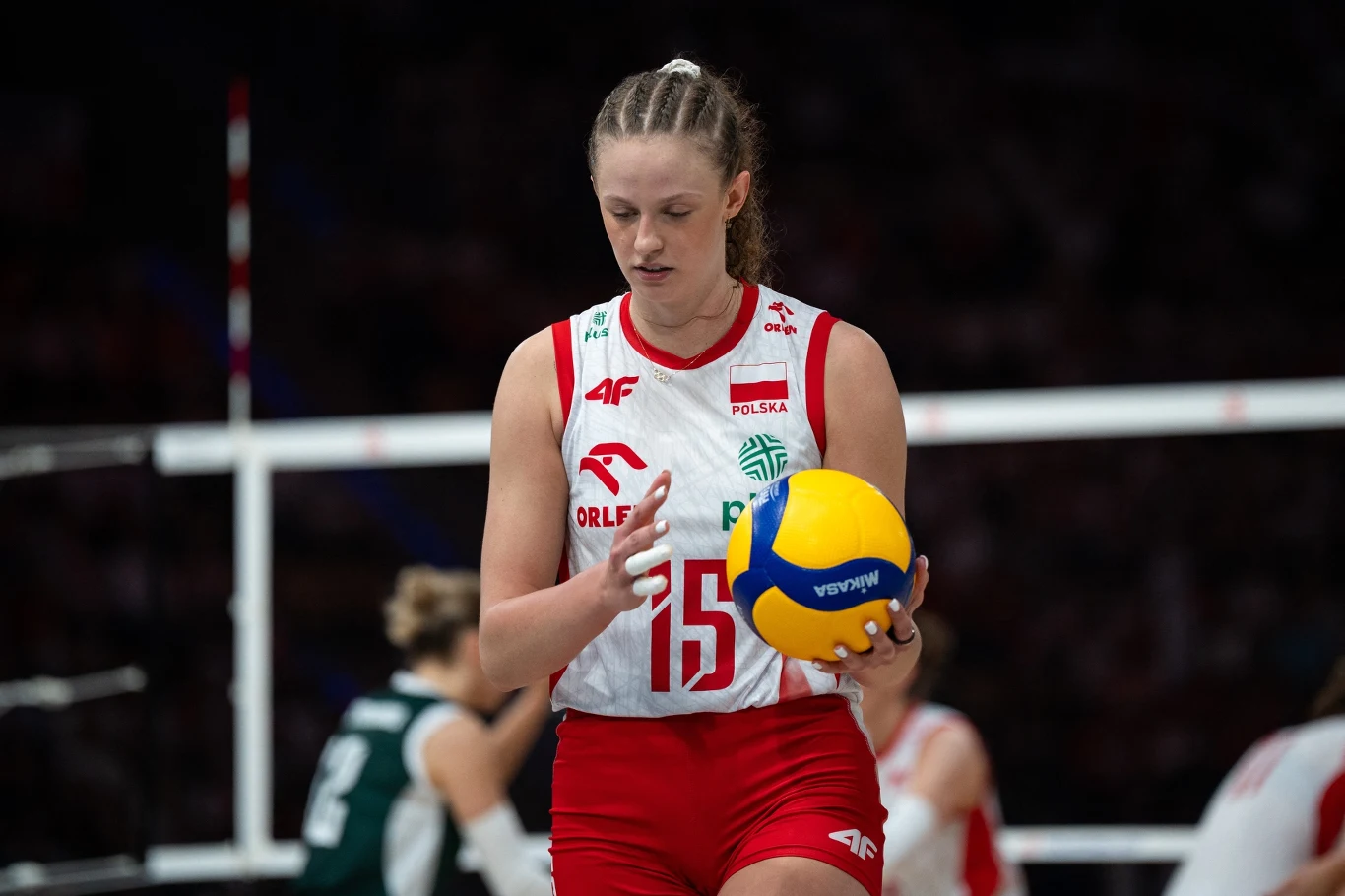 Martyna Czyrniańska Siatkarka w stroju reprezentacji Polski z numerem 15 trzyma piłkę do siatkówki na boisku, skupiona przed wykonaniem zagrania. W tle pozostali zawodnicy i elementy siatki.