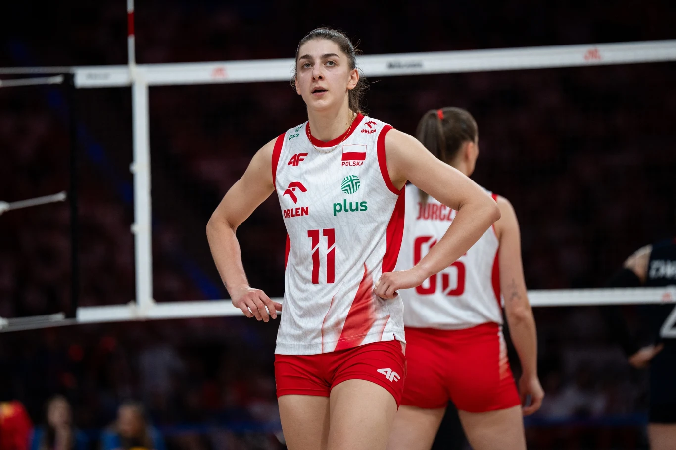 Zawodniczka reprezentacji Polski w siatkówce w biało-czerwonym stroju z numerem 11 na tle siatki, obok widoczna jest druga siatkarka z tej samej drużyny. Obiekt znajduje się na hali sportowej podczas meczu.