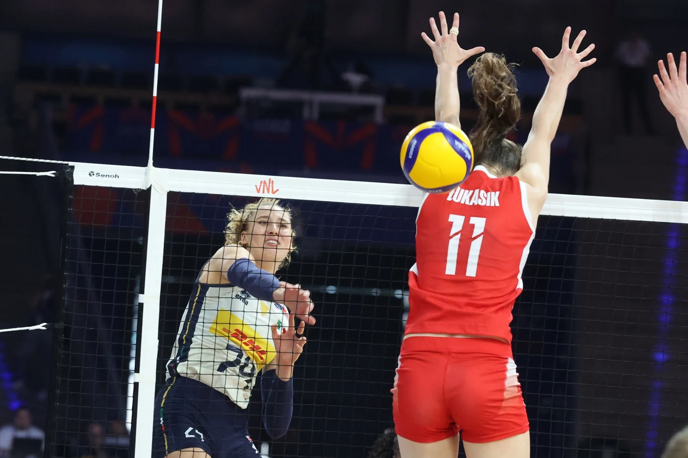 Martyna Łukasik (w bloku) Zawodniczka drużyny w granatowych spodenkach i jasnej koszulce wykonuje atak siatkarski na tle siatki, podczas gdy zawodniczka w czerwonym stroju z numerem 11 unosi ręce do bloku. Piłka znajduje się tuż nad siatką pomiędzy nimi.