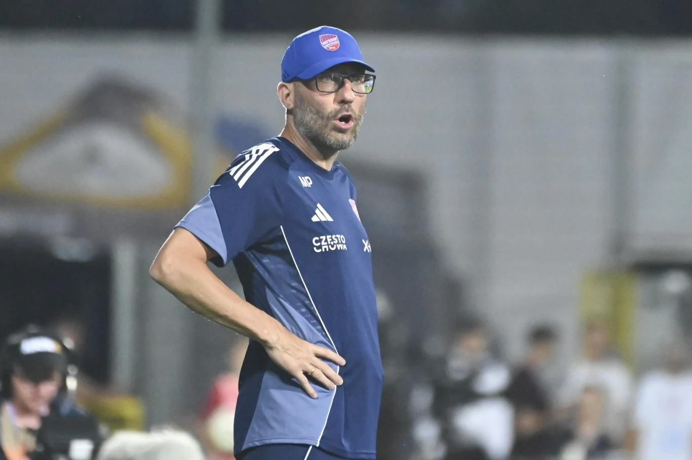 Marek Papszun Mężczyzna w sportowym stroju trenerskim, z czapką i okularami, stoi na boisku, w tle rozmyte postaci oraz elementy stadionu.
