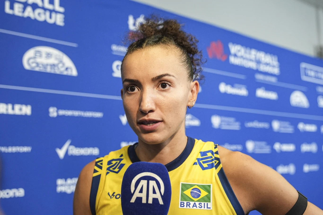Zawodniczka brazylijskiej reprezentacji siatkarskiej udziela wywiadu, ubrana w żółty strój sportowy z flagą Brazylii, w tle niebieska ściana z logotypami sponsorów i nazwą rozgrywek.