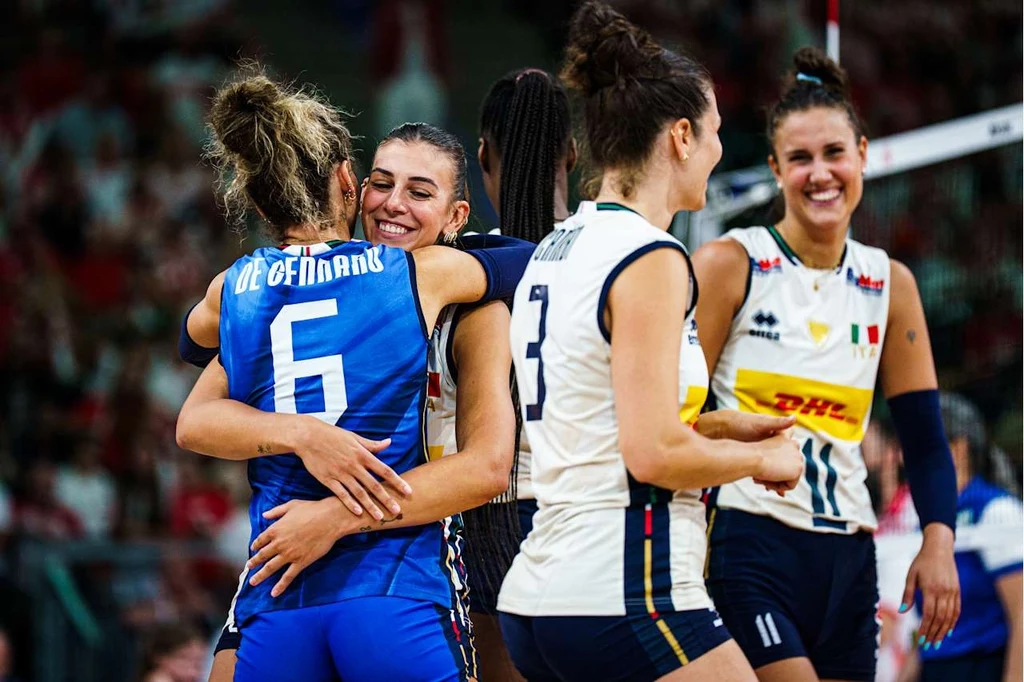 Włoskie siatkarki mogły świętować po zwycięstwie z Polską. Z prawej Anna Danesi Siatkarki w sportowych strojach celebrują zwycięstwo, obejmując się i uśmiechając; w tle rozmyta publiczność oraz siatka siatkarska.