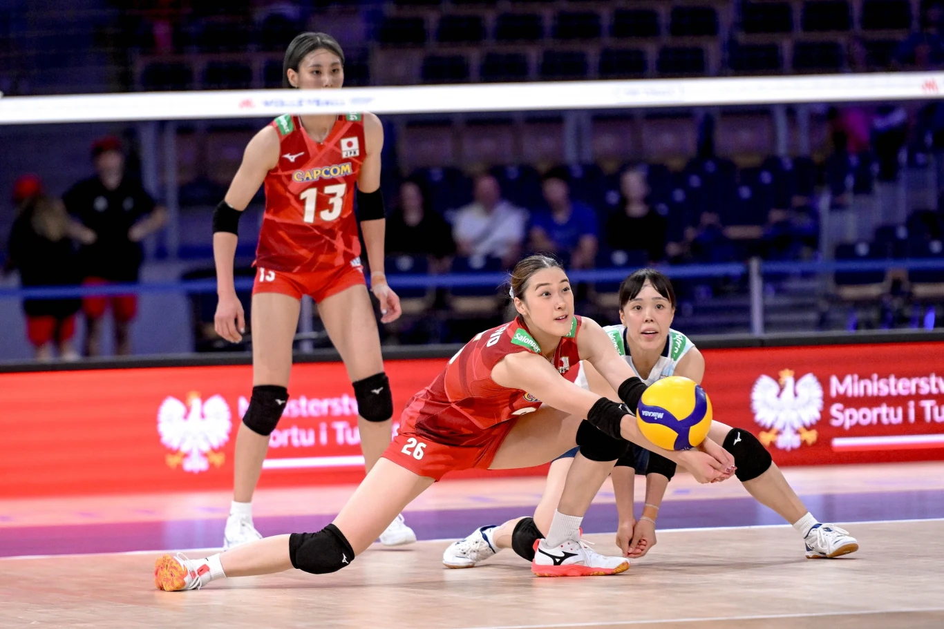 Zawodniczki drużyny Japonii ubrani w czerwone stroje sportowe podczas dynamicznej obrony w meczu siatkówki, jedna z nich wykonuje odbiór piłki w niskiej pozycji, a pozostałe dwie są przygotowane do kolejnej akcji.