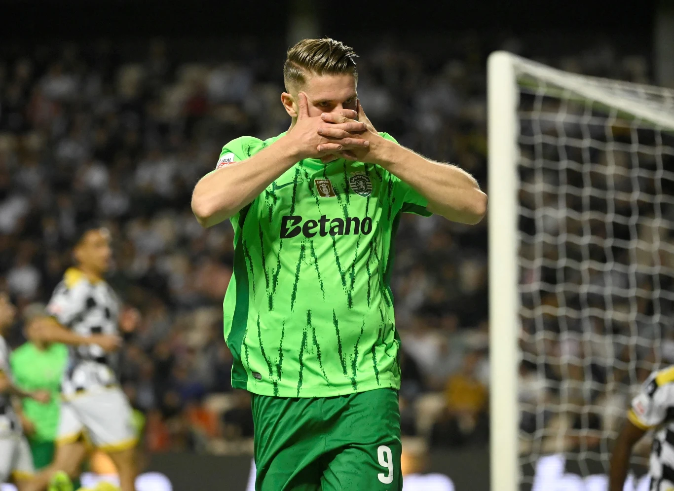 Viktor Gyökeres Piłkarz w zielonej koszulce z numerem 9 wykonuje gest dłońmi na twarzy podczas meczu piłki nożnej, w tle widoczni są inni zawodnicy oraz widownia.