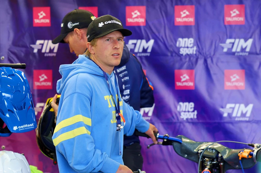 Andrzej Lebiediew Młody mężczyzna w sportowej bluzie i czapce z daszkiem stoi obok motocykla, w otoczeniu sprzętu wyścigowego, na tle fioletowej ścianki z logotypami sponsorów i organizatorów zawodów.