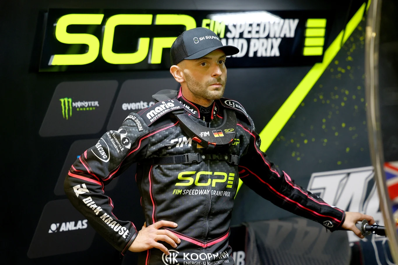 Zawodnik żużlowy ubrany w kombinezon wyścigowy z widocznymi logotypami sponsorów oraz napisem SGP FIM Speedway Grand Prix, stojący przy stanowisku w otoczeniu banerów reklamowych i sprzętu mechanicznego.