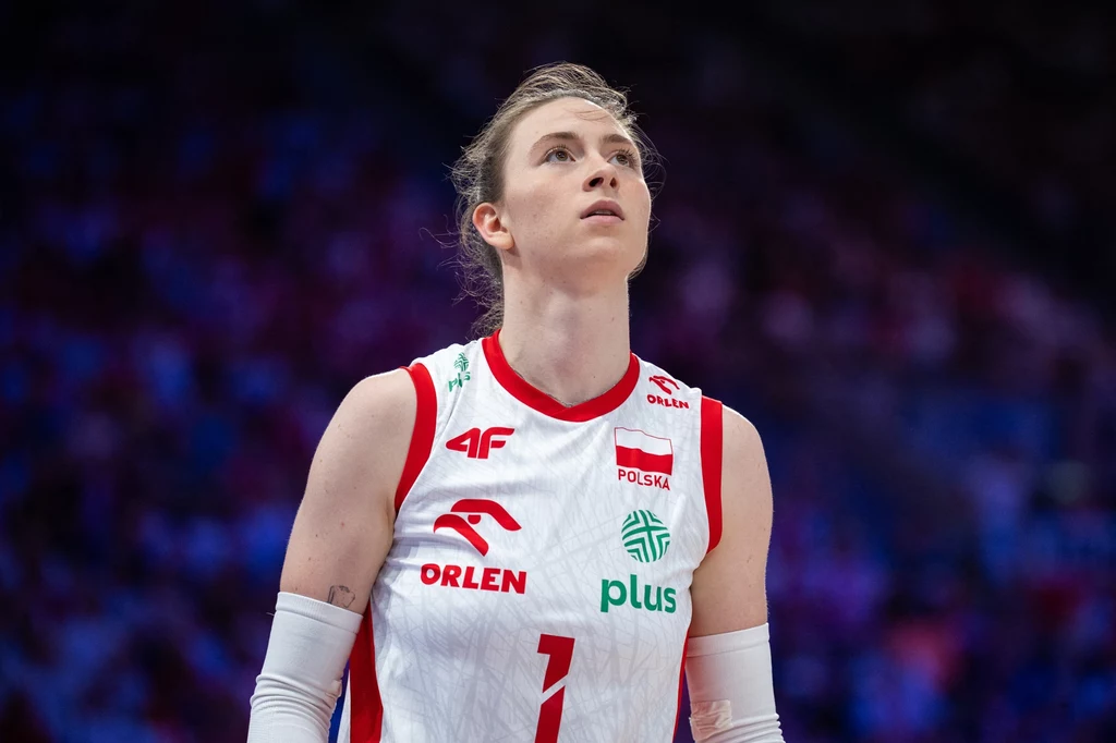 Aleksandra Gryka Zawodniczka w stroju reprezentacji Polski do siatkówki, skupiona na tle rozmytego stadionu, wyraźnie przygotowująca się do kolejnej akcji w grze.