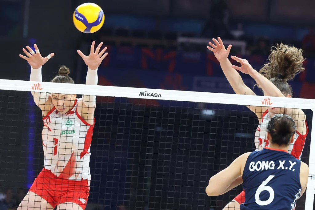 Aleksandra Gryka skacze do bloku Dwie zawodniczki w czerwono-białych strojach blokują atak rywalki w granatowym stroju po drugiej stronie siatki, w tle piłka siatkowa unosząca się nad siatką podczas dynamicznej akcji sportowej.