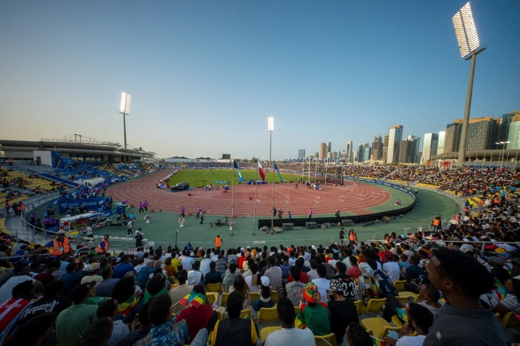 Stadion lekkoatletyczny wypełniony kibicami na trybunach, zielona murawa wewnątrz bieżni, sportowcy oraz obsługa zawodów rozmieszczeni na różnych częściach areny, wysokie oświetlenie stadionowe, w tle panorama nowoczesnych budynków.