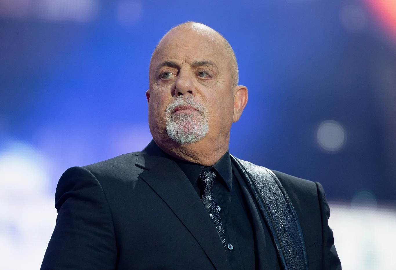 Billy Joel podzielił się wrażeniami z legendarnego Woodstocku Łysy mężczyzna w średnim wieku, ubrany w ciemny garnitur i krawat, patrzy w bok na tle rozmytego, kolorowego światła scenicznego. Na ramieniu widoczny pasek od gitary.
