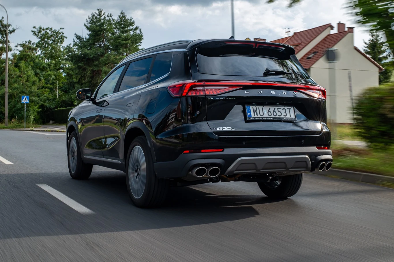 Chery Tiggo 8 pokazało w teście pełen wachlarz swoich zalet. Nie ustrzegło się wad Czarny SUV marki Chery Tiggo 8 z widoczną rejestracją porusza się po miejskiej ulicy w otoczeniu zieleni oraz budynków mieszkalnych, z tyłu widoczne są cztery końcówki układu wydechowego oraz charakterystyczne tylne lampy