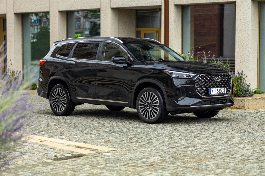 Czarny nowoczesny SUV zaparkowany na brukowanym placu przed budynkiem o prostych, betonowych elewacjach i dużych oknach.