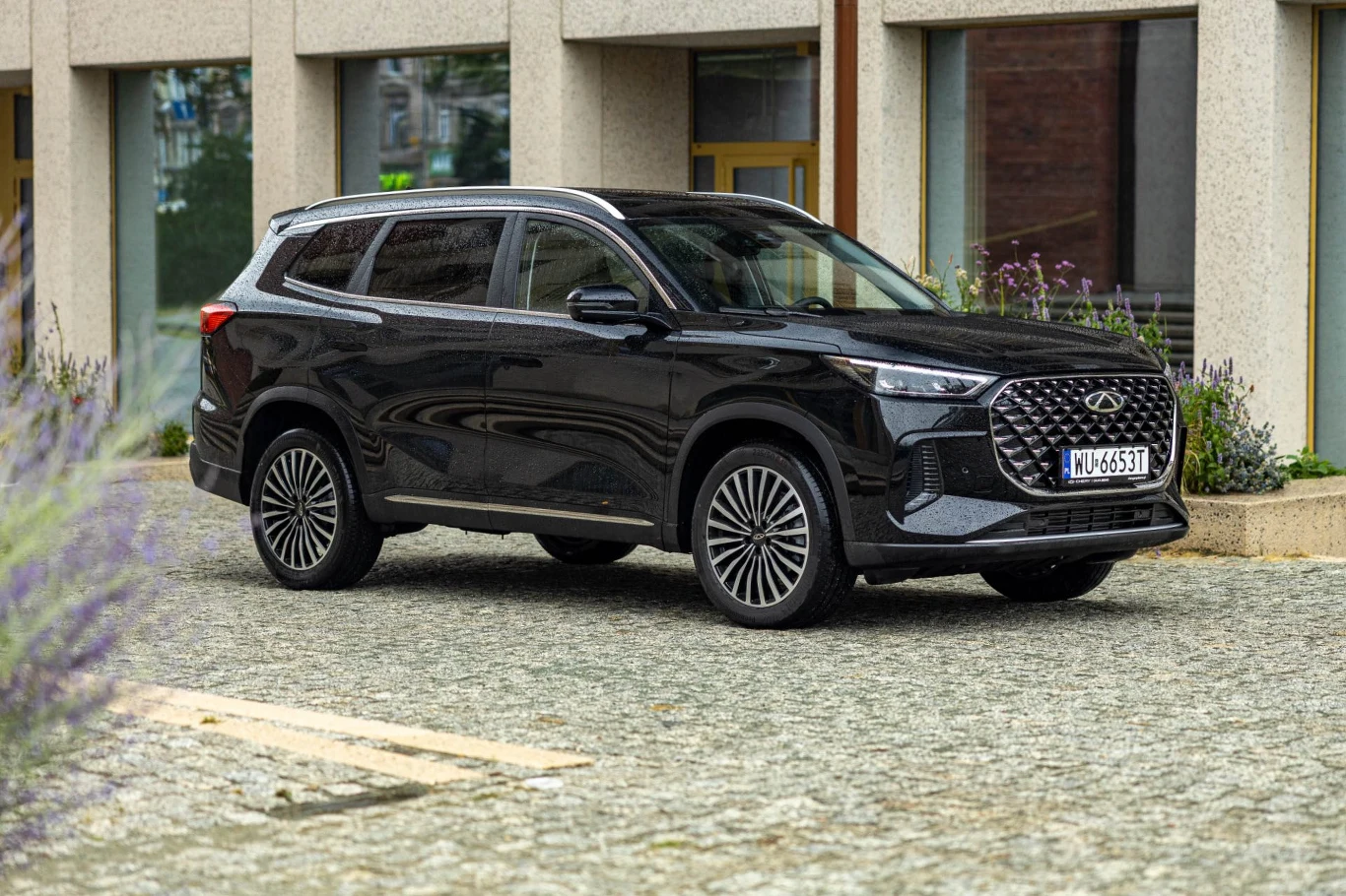 Chery Tiggo 8 w Polsce dostępny jest wyłącznie w wersji 7-miejscowej Czarny nowoczesny SUV zaparkowany na brukowanym placu przed budynkiem o prostych, betonowych elewacjach i dużych oknach.