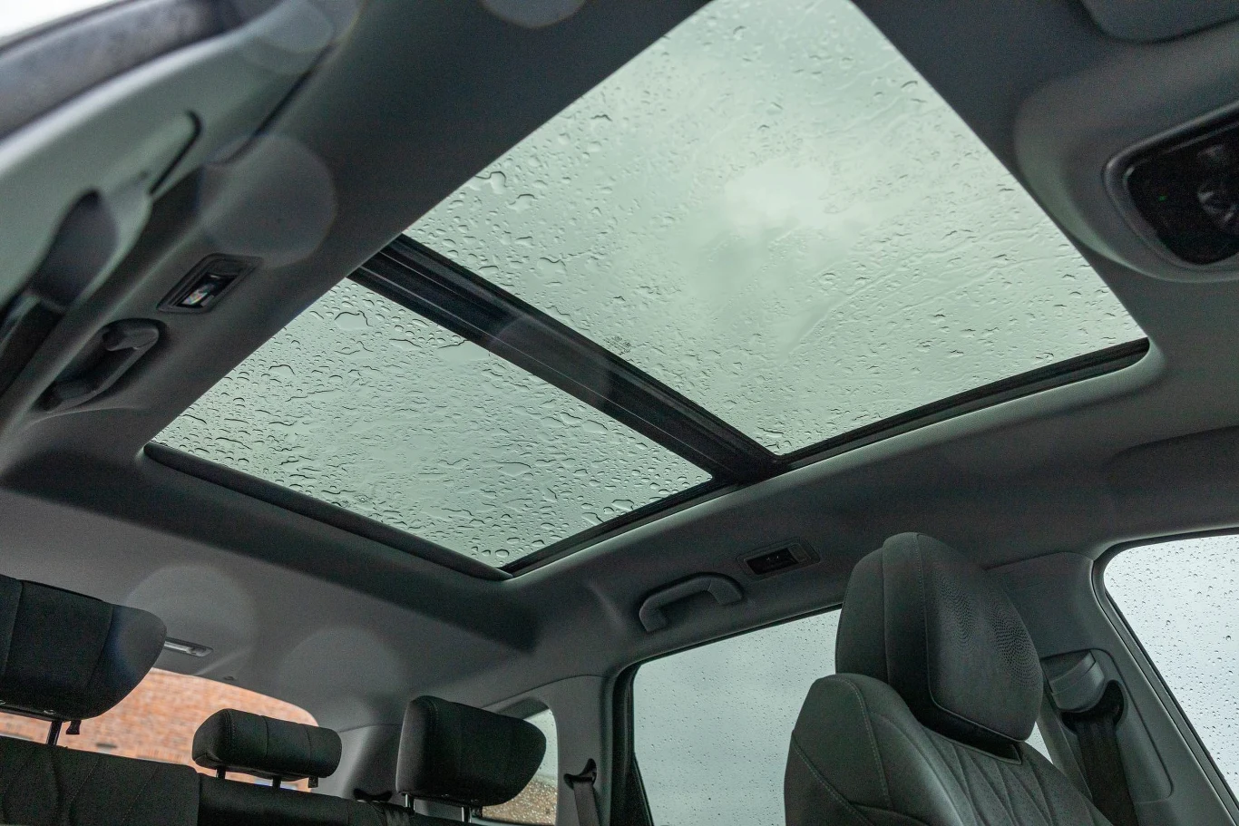 Wielki szyberdach ma elektrycznie sterowaną przesłonę. Reaguje na komendy głosowe Wnętrze nowoczesnego samochodu z widocznym panoramicznym dachem szklanym pokrytym kroplami deszczu, z przodu dwa skórzane fotele oraz część tylnej kanapy.