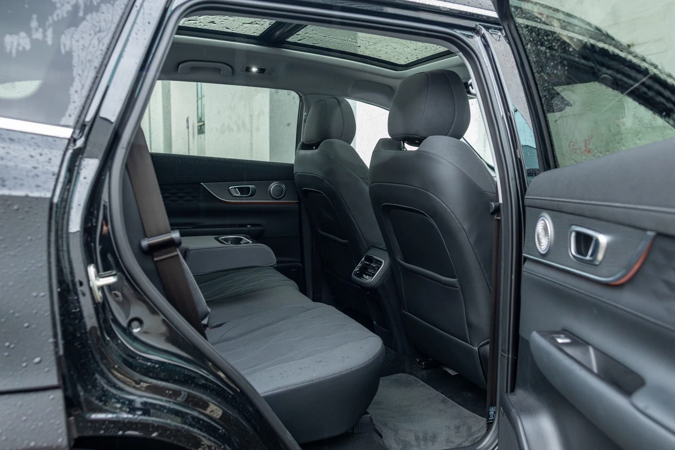 W drugim rzędzie jest naprawdę przestronnie. Kanapę można przesunąć o 40 cm Wnętrze samochodu osobowego z widocznym tylnym siedzeniem, otwartymi drzwiami i panoramicznym dachem, tapicerka w odcieniach szarości, na zewnątrz wyraźne ślady deszczu na karoserii.