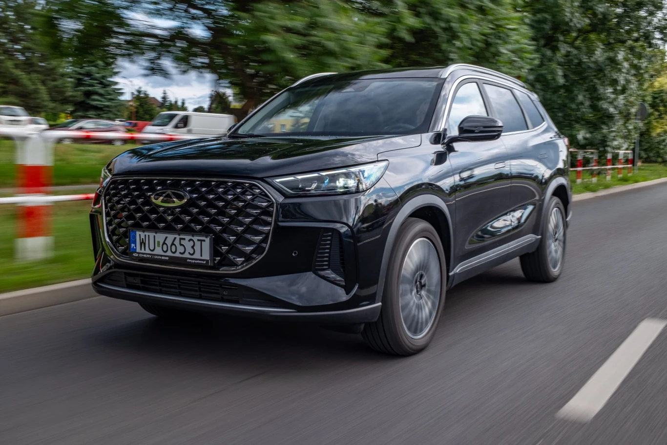 Chery Tiggo 8 wyjeżdża z salonu na 19-calowych obręczach Czarny samochód typu SUV marki Chery Tiggo 8 jedzie po asfaltowej drodze w otoczeniu zielonych drzew i innych pojazdów widocznych w tle.