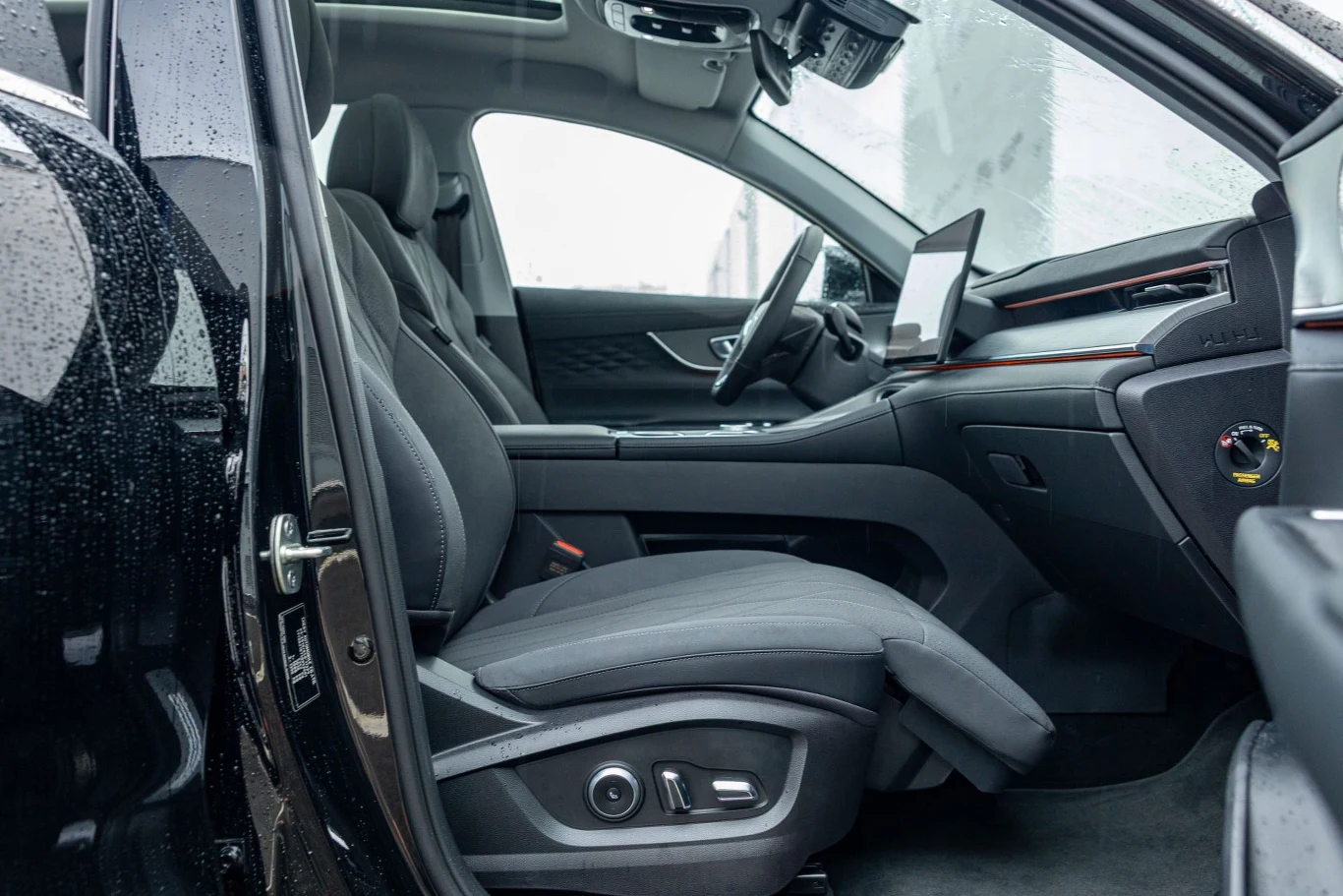 "Fotel królowej" – ma rozkładany podnóżek, jest podgrzewany i wentylowany, a dodatkowo jako jedyny został wyposażony w funkcję masażu Nowoczesne, przestronne wnętrze samochodu z czytelnym wyświetlaczem na desce rozdzielczej, komfortowymi fotelami i detalami z eleganckiego materiału, widok z otwartymi drzwiami podczas deszczowej pogody.