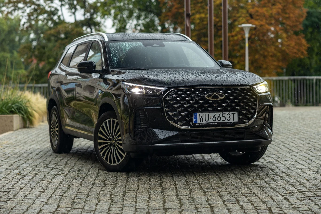 Przednią część nadwozia zdominował wielki grill z błyszczącymi wstawkami Czarny nowoczesny SUV o charakterystycznym dużym grillu na froncie, stojący na brukowanej nawierzchni, otoczony zielenią oraz jesiennymi drzewami w tle, lekko mokry karoserii od deszczu.