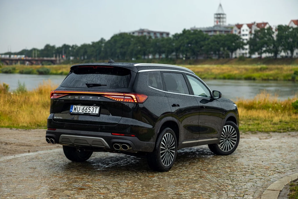 Czarny SUV Geely Okavango zaparkowany na brukowanej nawierzchni, w tle widać rzekę, trawę oraz zabudowania z białymi elewacjami i wieżą.