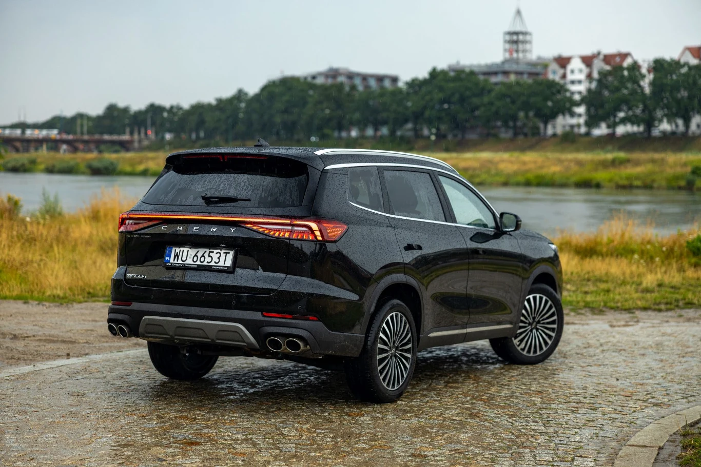 Wielki samochód za sensowne pieniądze. Tiggo 8 warto wpisać na listę aut do sprawdzenia Czarny SUV Geely Okavango zaparkowany na brukowanej nawierzchni, w tle widać rzekę, trawę oraz zabudowania z białymi elewacjami i wieżą.