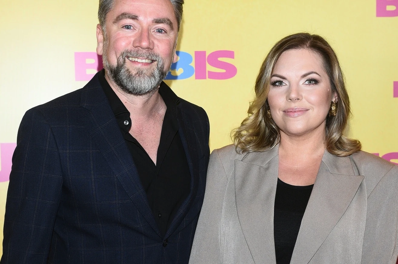 Kuba Badach i Aleksandra Kwaśniewska podzielili się radosną nowiną Dwie uśmiechnięte osoby, mężczyzna w ciemnej marynarce i kobieta w jasnym płaszczu stojące na tle żółtej ścianki z kolorowym napisem BIS.