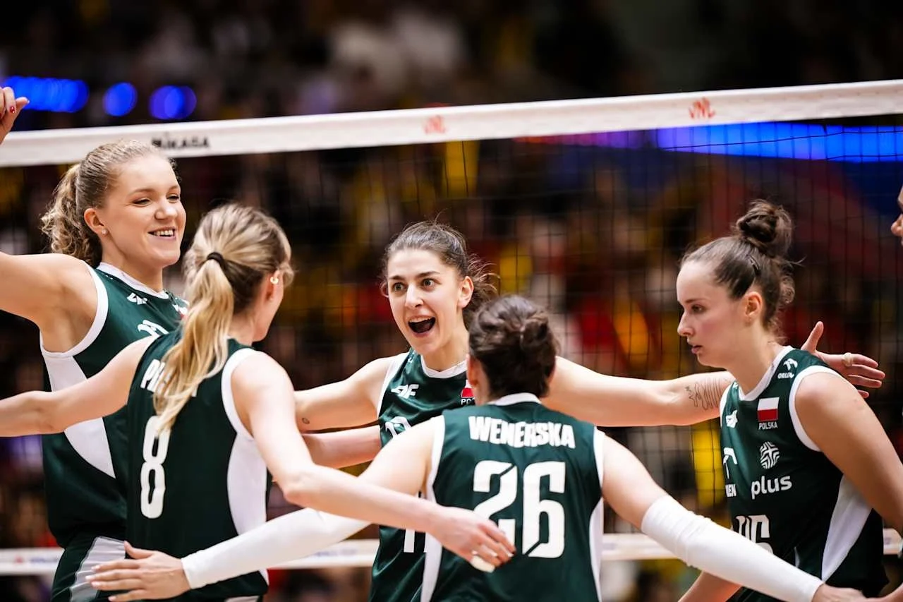 Grupa siatkarek w zielonych strojach z numerami i napisem Polska na piersi, celebrujących punkt przy siatce na hali sportowej. Zawodniczki są uśmiechnięte, z uniesionymi rękami, w tle rozmyte trybuny.