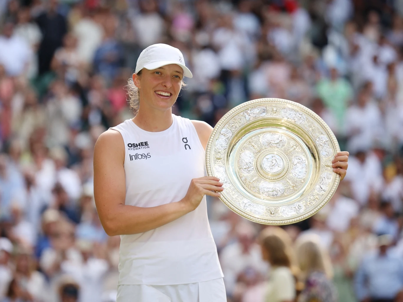 Iga Świątek po triumfie na Wimbledonie Uśmiechnięta tenisistka w białym stroju sportowym trzyma złotą, okrągłą trofeum na tle zamazanych sylwetek widzów na trybunach.
