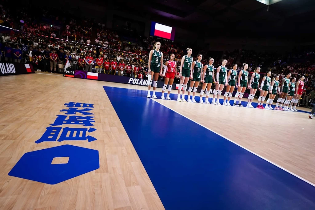 Liga Narodów siatkarek. Polska zagra z Japonią Zespół siatkarek w zielonych strojach ustawionych w szeregu na boisku tuż przed rozpoczęciem meczu, po lewej stronie widoczne fragmenty logotypów i napisy na parkiecie, w tle tłum kibiców i polska flaga na ekranie.
