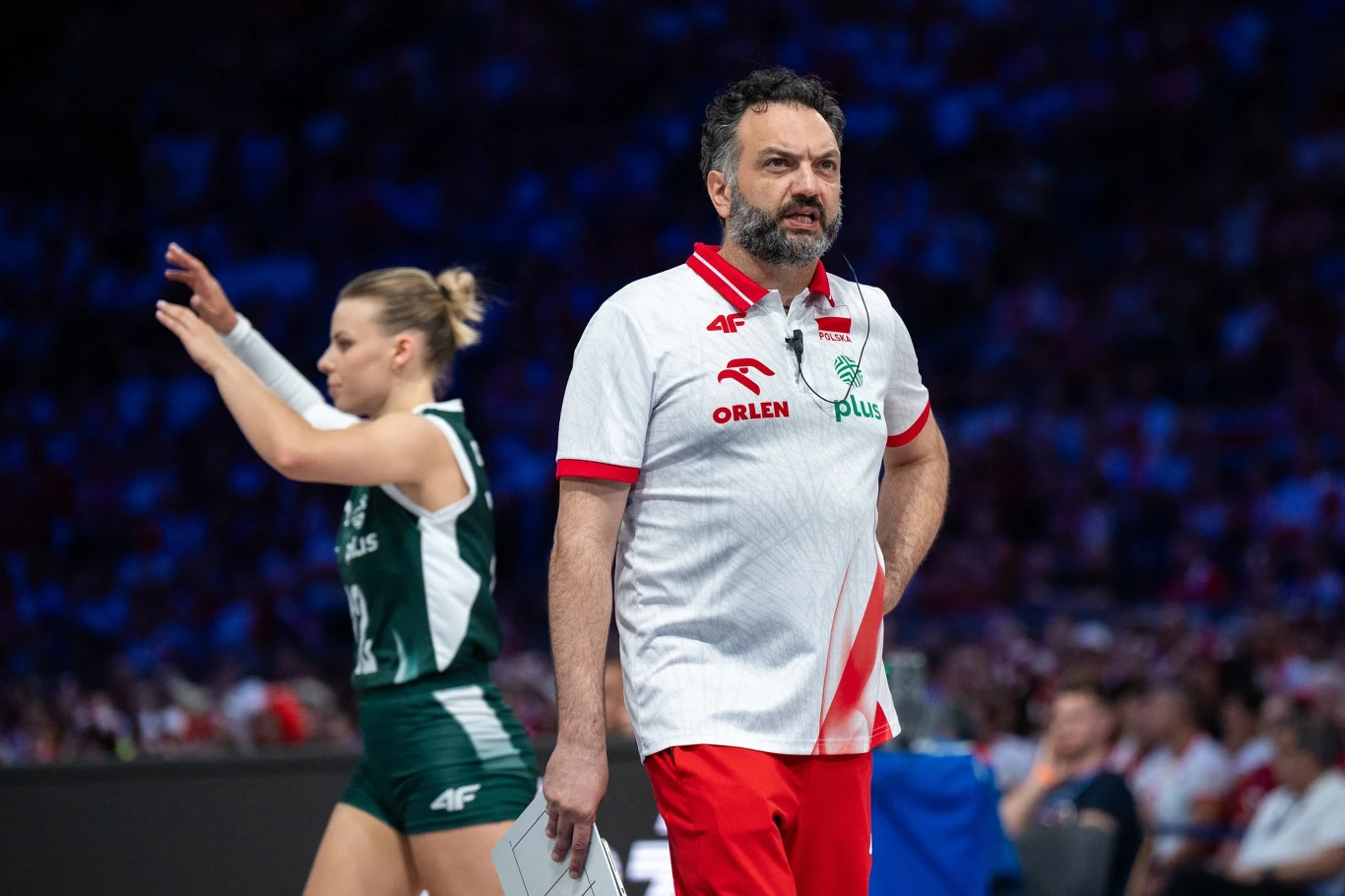 Trener drużyny siatkarskiej w stroju sportowym stoi na hali podczas meczu, w tle siatkarka w zielonym stroju wykonuje gest ręką, otoczenie wypełnia rozmyty tłum kibiców.