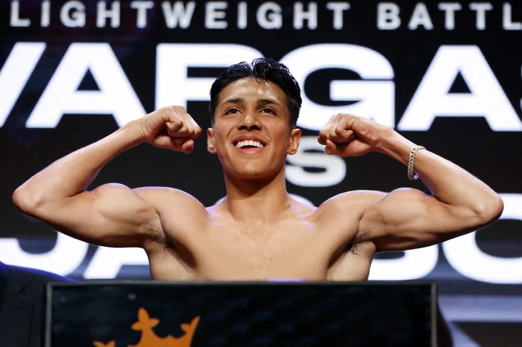 Emiliano Vargas to jeden z najbardziej utalentowanych bokserów w swojej kategorii wagowej Młody mężczyzna z uśmiechem prezentuje napięte bicepsy, stojąc bez koszulki na tle tablicy z napisem i światłami, prawdopodobnie podczas oficjalnego ważenia przed walką sportową.