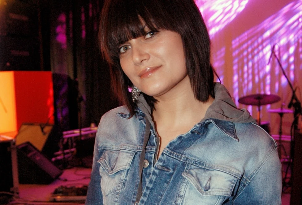 Sylwia Wiśniewska podbiła listy przebojów utworem "12 łez" Młoda kobieta w dżinsowej kurtce stoi na tle kolorowego, podświetlonego ekranu scenicznym, w tle widoczne są instrumenty muzyczne i sprzęt nagłośnieniowy.