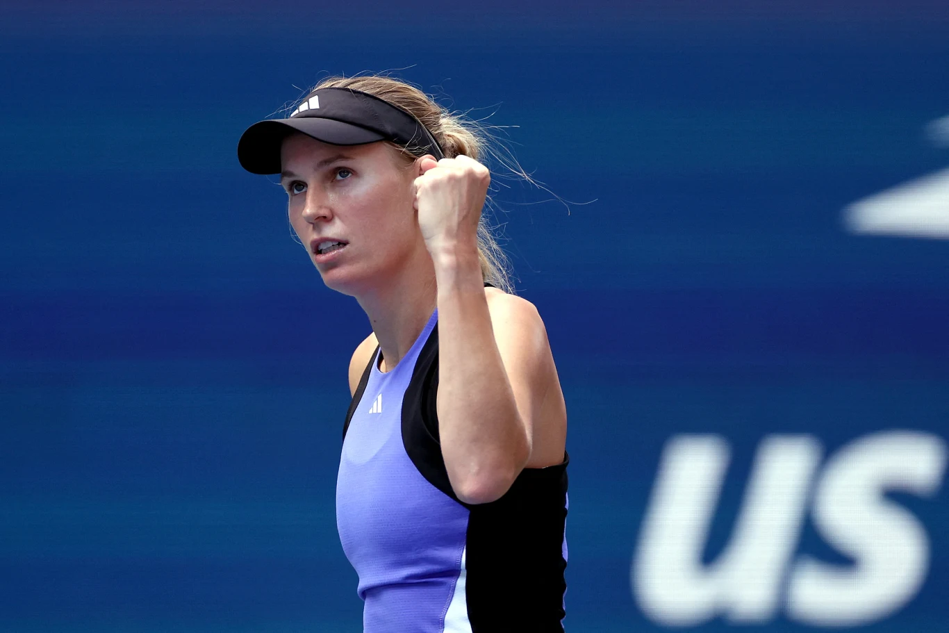 Tenisistka w niebieskim stroju sportowym, z czapką na głowie, zaciśniętą pięścią okazuje emocje na tle niebieskiego kortu i częściowo widocznego logo turnieju.