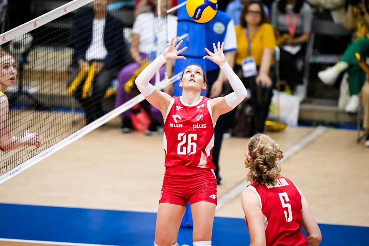 Zawodniczka w czerwonym stroju z numerem 26 szykuje się do odbicia piłki podczas meczu siatkówki, obok niej inna zawodniczka z numerem 5, w tle widoczna siatka, sędzia oraz publiczność.