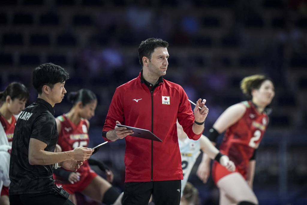 Ferhat Akbas - trener reprezentacji Japonii Trener reprezentacji Japonii w czerwonej bluzie prowadzi odprawę na tle swoich zawodniczek ubranek w czerwone stroje siatkarskie podczas meczu siatkówki, skupienie na twarzach sportowców, dynamiczna atmosfera rozgrywki.