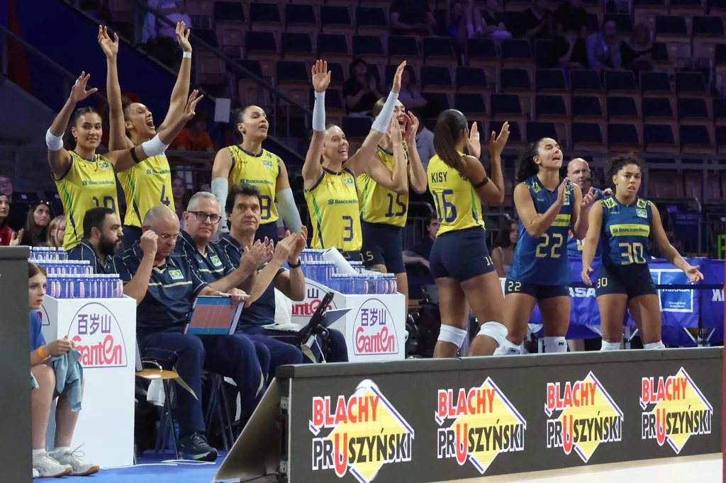 Siatkarki reprezentacji Brazylii Zawodniczki drużyny siatkówki w żółto-niebieskich strojach oraz sztab trenerski na ławce rezerwowych w hali sportowej, część siatkarek cieszy się i unosi ręce w geście radości, trybuny w tle są puste