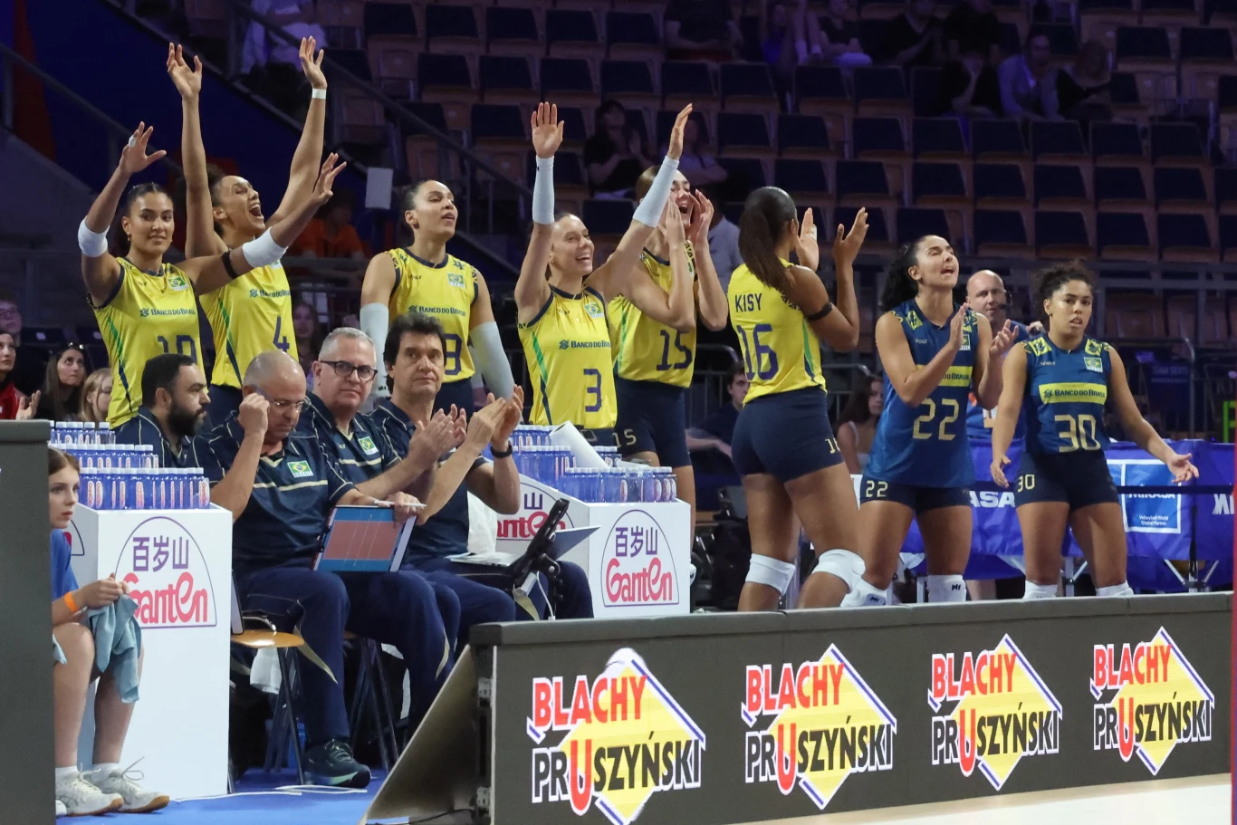 Zawodniczki drużyny siatkówki w żółto-niebieskich strojach oraz sztab trenerski na ławce rezerwowych w hali sportowej, część siatkarek cieszy się i unosi ręce w geście radości, trybuny w tle są puste