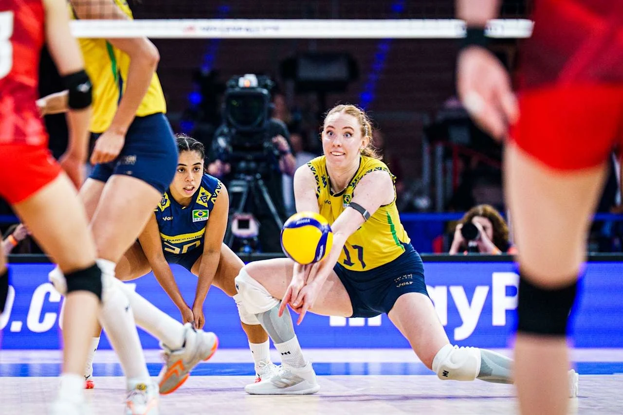 Zawodniczka w żółto-niebieskim stroju odbiera piłkę podczas dynamicznej akcji na meczu siatkówki, w tle widoczne inne zawodniczki oraz elementy hali sportowej.