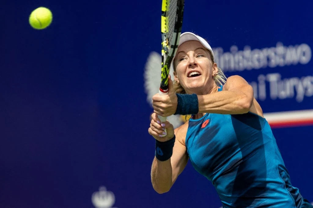 Tenisistka w niebieskiej sportowej sukience odbija piłkę tenisową rakietą podczas meczu na korcie na świeżym powietrzu, w tle widoczne nieostre logo Ministerstwa Sportu i Turystyki.