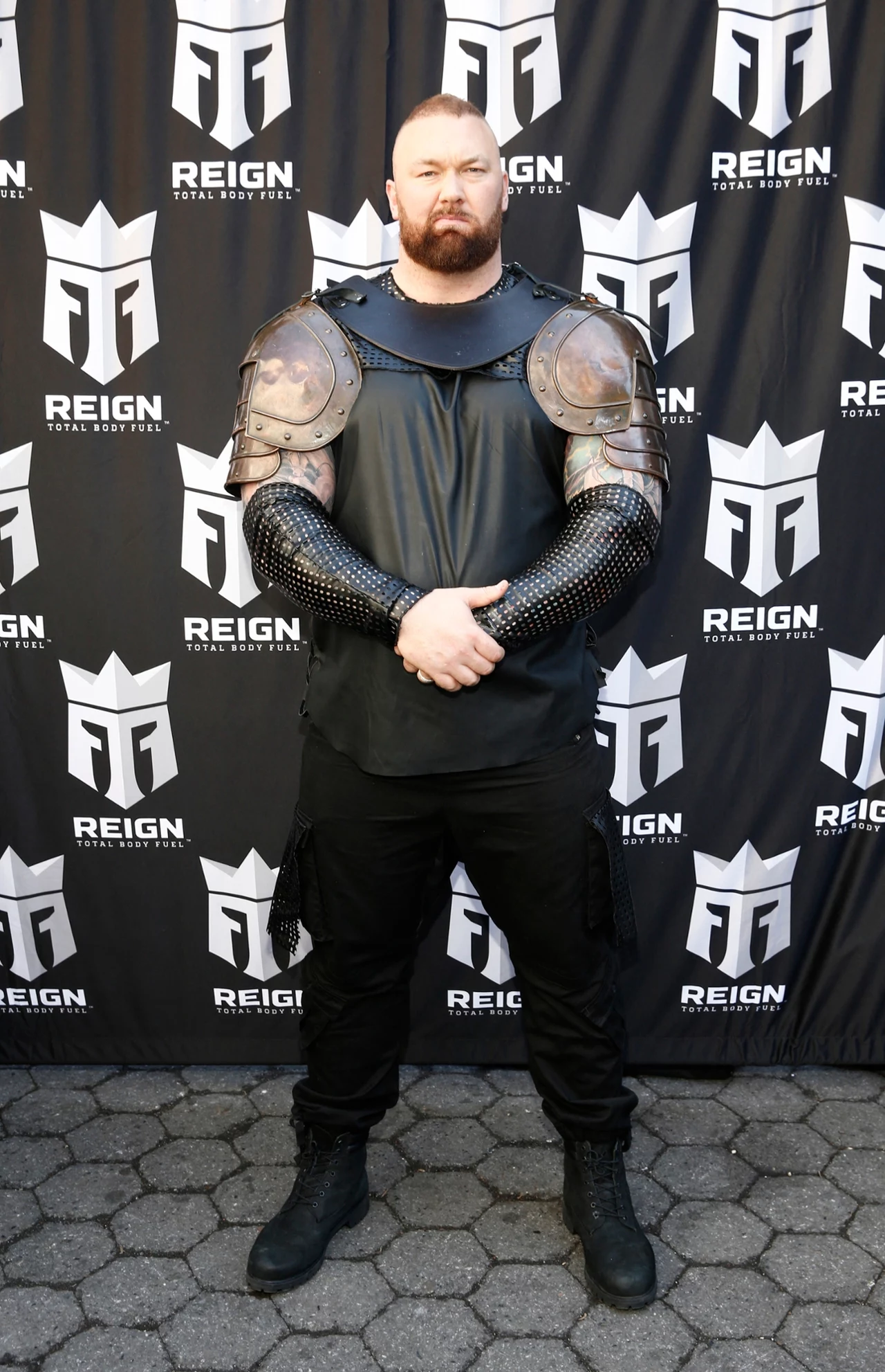 Hafthor Julius Bjornsson Mężczyzna o muskularnej sylwetce w średniowiecznym stroju wojownika, z kolczugą na ramionach i żelaznymi naramiennikami, stoi na tle ściany z logo Reign Total Body Fuel.