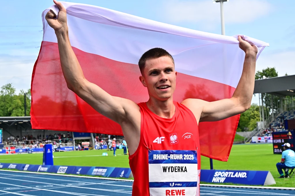 Lekkoatleta w czerwonym stroju sportowym unosi nad głową biało-czerwoną flagę Polski stojąc na niebieskiej bieżni stadionu, w tle widoczne są trybuny i inni sportowcy.