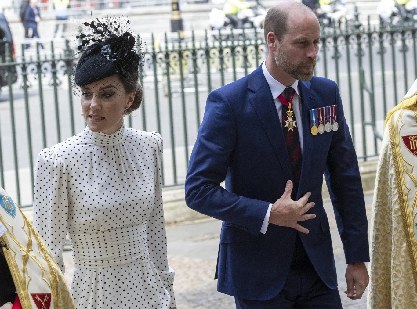 Księżna Kate i książę William od tygodnia przebywają na urlopie Elegancko ubrana kobieta w białej sukience w czarne grochy z ozdobnym kapeluszem oraz mężczyzna w granatowym garniturze z medalami na piersi, idący razem obok siebie w otoczeniu osób w ceremonialnych szatach, za nimi widoczny ogrodzony teren miejskiej ...